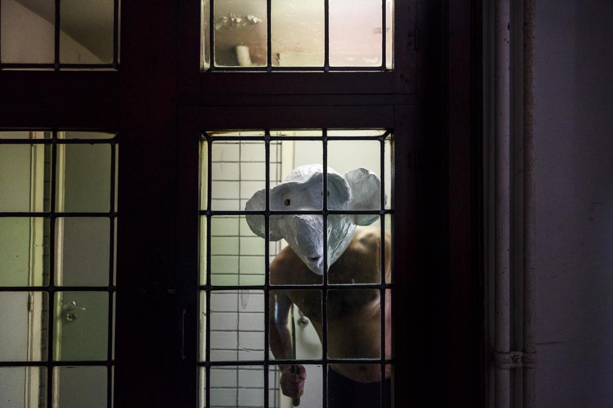 Man met wit masker en ontbloot bovenlijf achter huisraam bij nacht