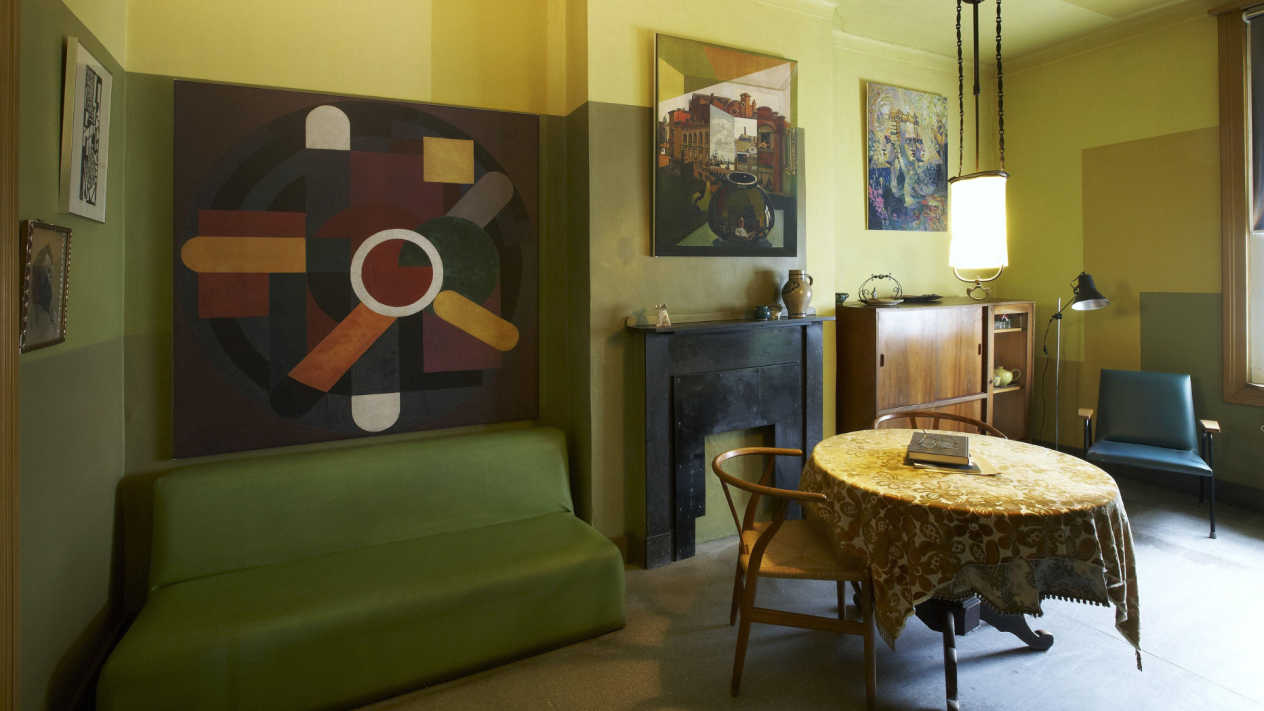 Woonkamer in de flat met groene sofabank, rond tafeltje met twee stoelen, een fauteuil met staanlamp ernaast. De muren zijn geschilderd in geometrische stroken in geeltinten en er hangen kunstwerken aan de muren.