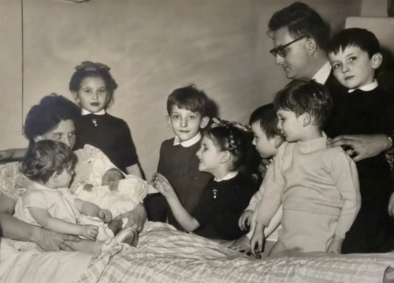 Een moeder zit rechtop in bed met een pasgeboren baby in haar armen. De 7 andere kinderen en de vader staan errond.