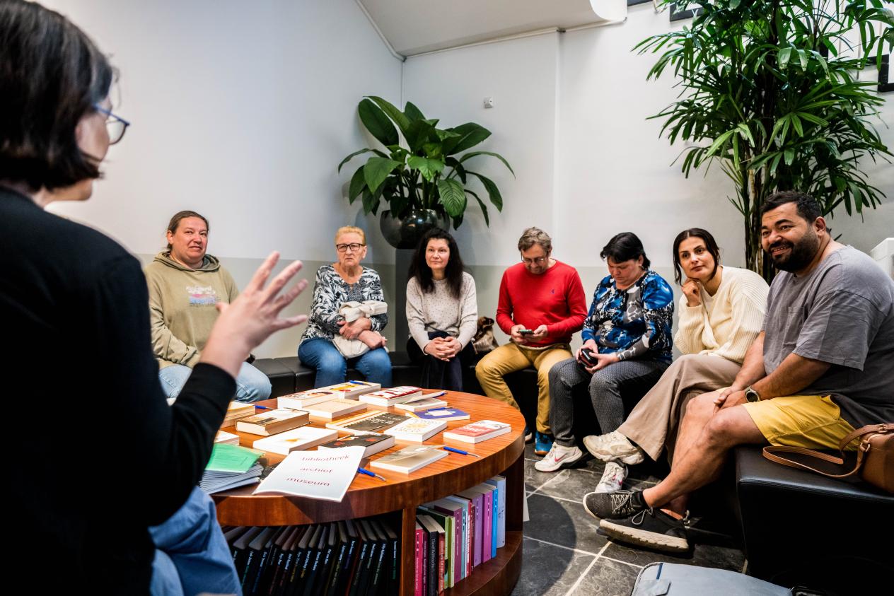 Een groep volwassenen zit in een kring in de leeshoek en luistert naar de uitleg van de gids