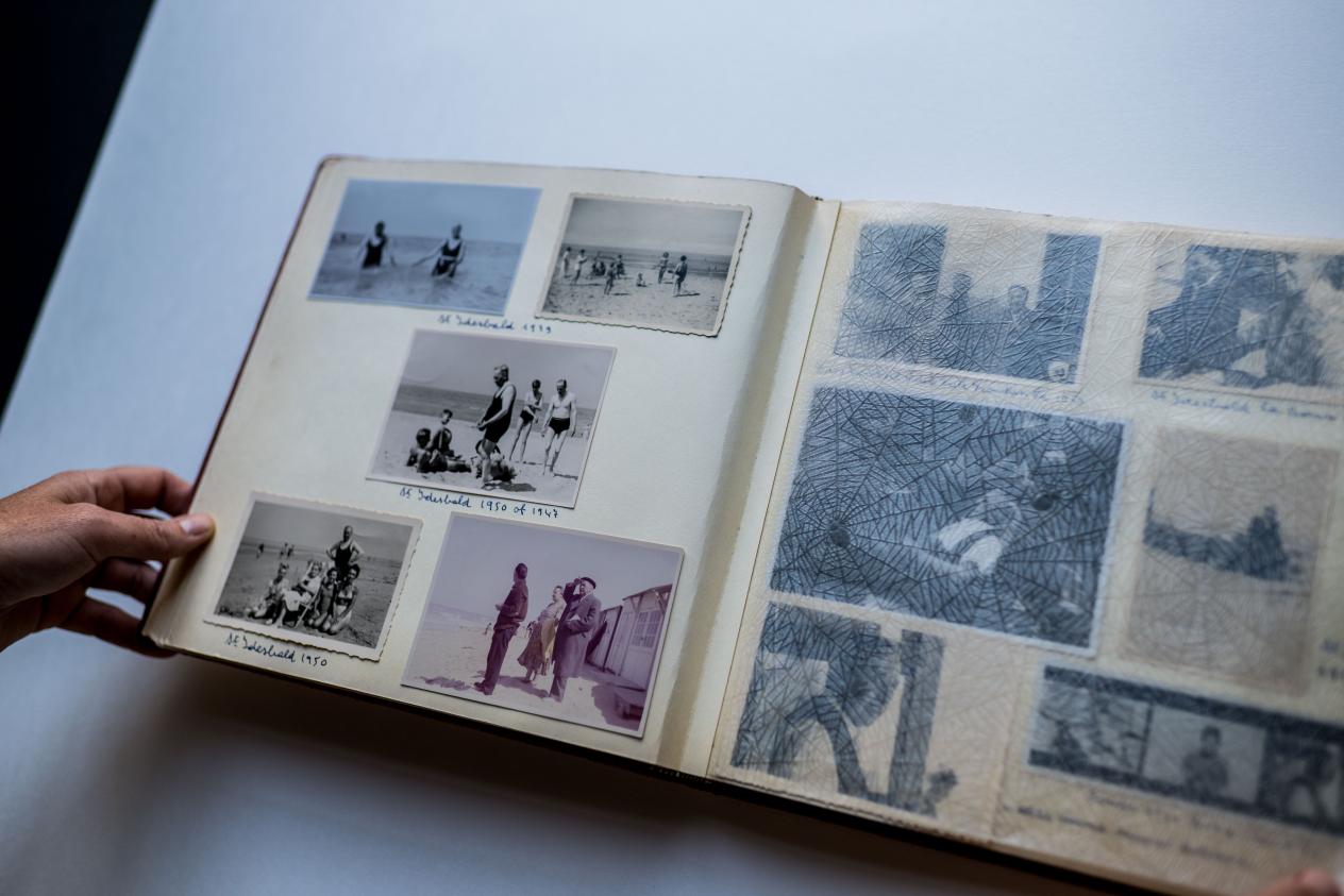 Een hand houdt een fotoalbum open dat op de tafel ligt, met verschillende zwart-witfoto’s van een strand.