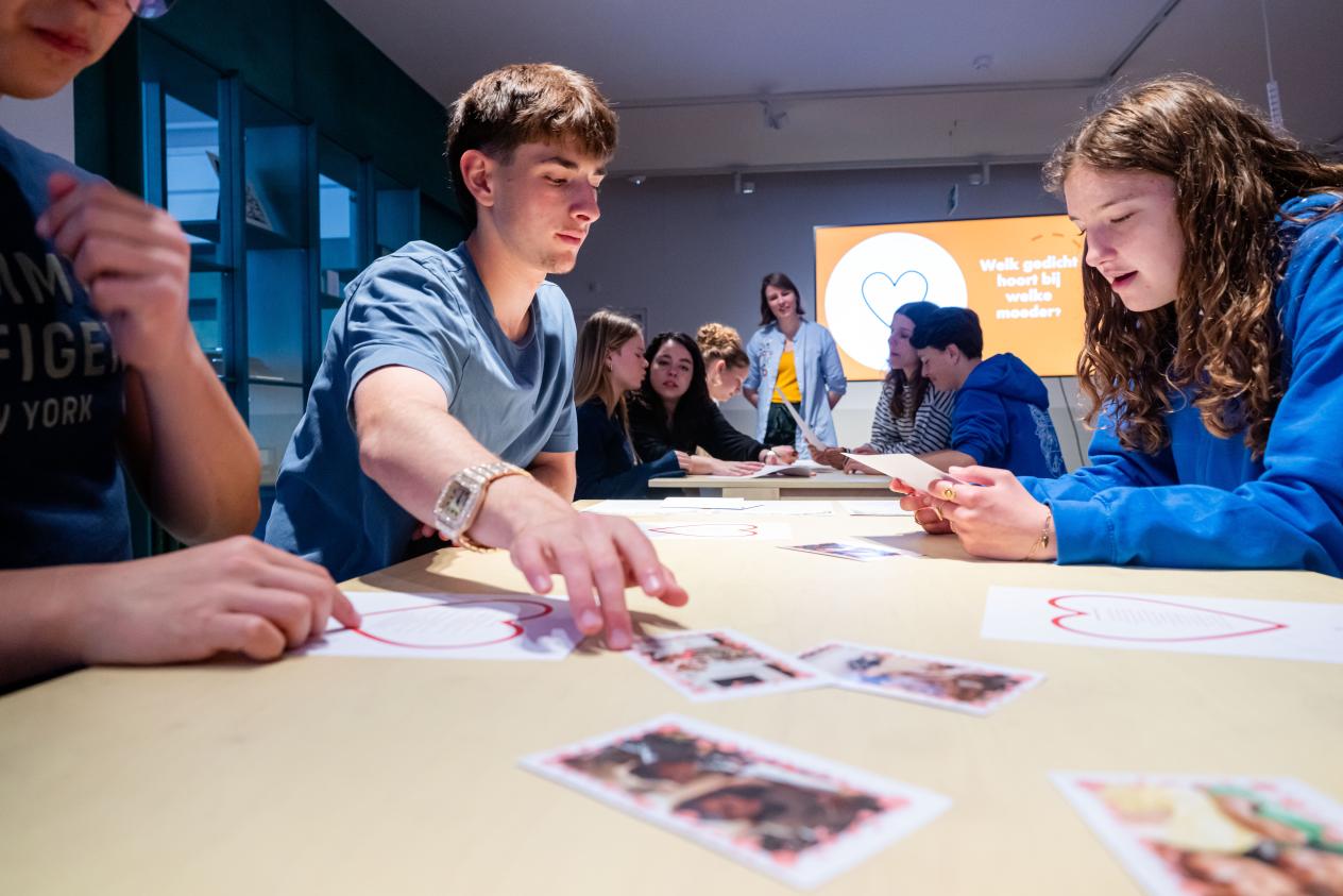 Drie leerlingen zitten aan een tafel waarop enkele foto's van moeders liggen en bespreken deze foto's. Op de achtergrond een tweede tafel met leerlingen rond en de workshopbegeleider die erbij staat.