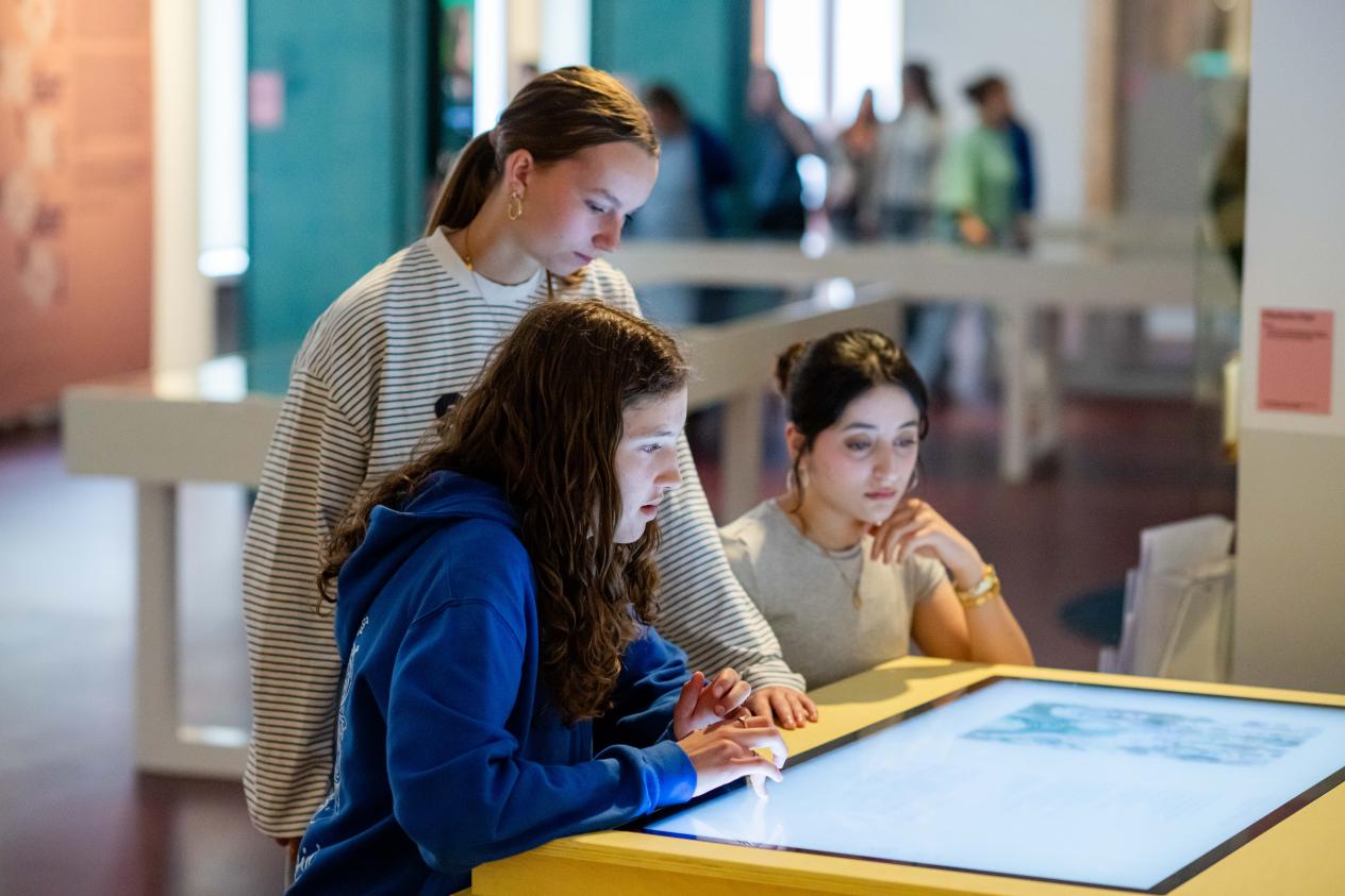 Drie meisjes zitten rond een touchscreen in de thema-expo Moeders en beantwoorden de vragen op het scherm.