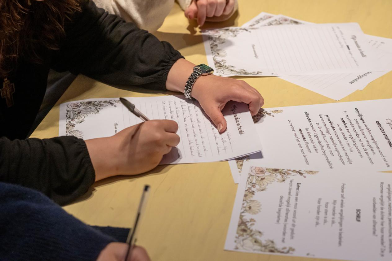 Op een tafel liggen een aantal voorbeelden van gedichten voor moeders. Naast de gedichten wordt een blad beschreven met een nieuw moederdaggedicht, enkel de handen die het blad en de pen vasthouden zijn zichtbaar).