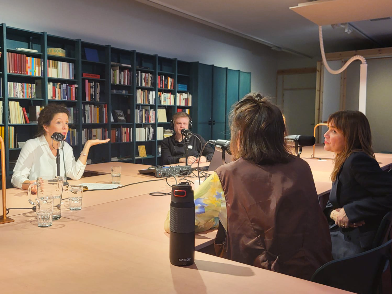 3 vrouwen en een man zijn in gesprek aan de tafels in de leeszaal van het Letterenhuis, op de tafels liggen schriften met notities en afgedrukte documenten. Het gezelschap praat in de microfoons die deel uitmaken van de podcastset.