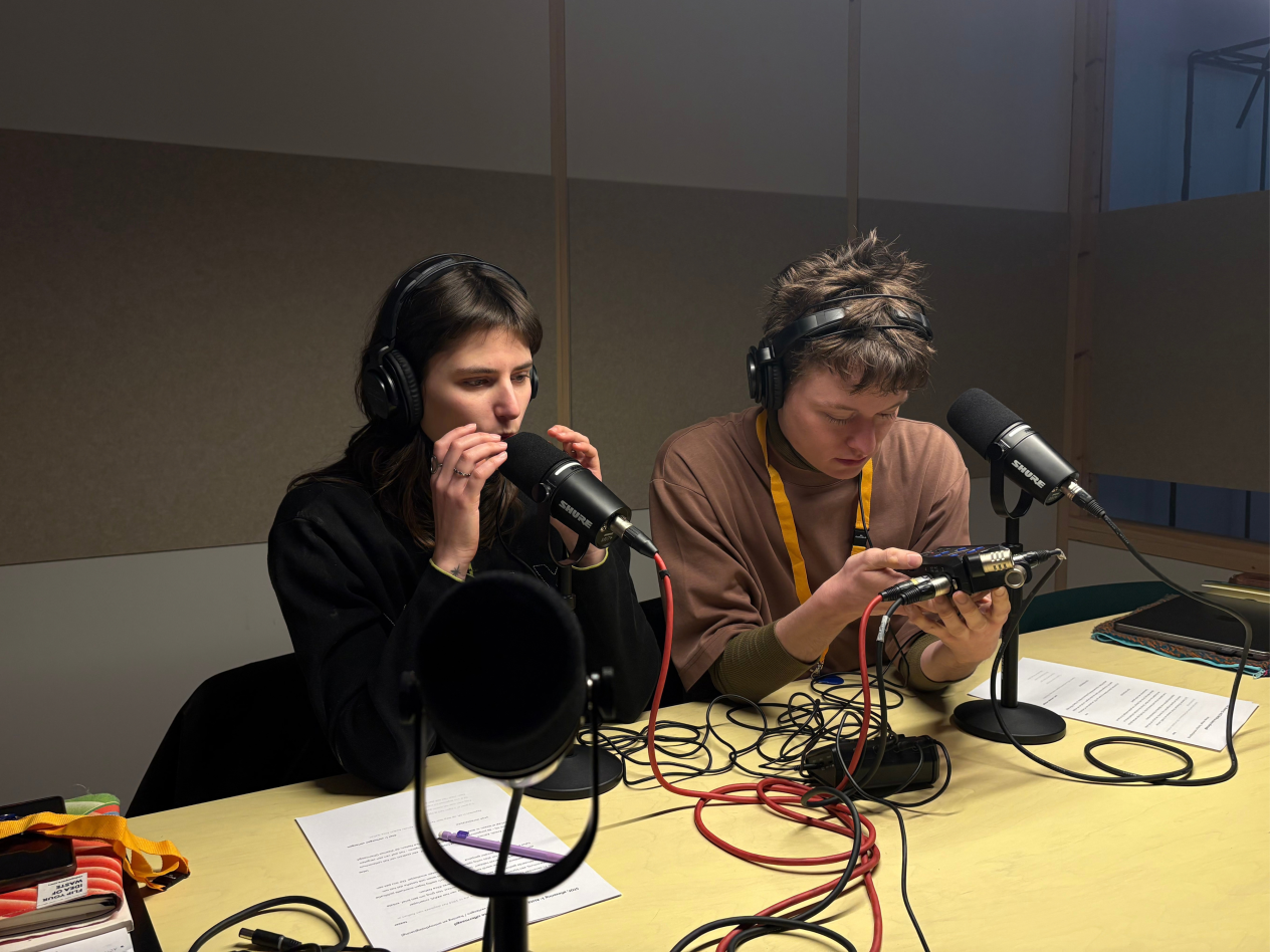 Een jonge man en een jonge dame zitten samen aan een tafel in de vergaderruimte met voor hen twee podcastmicrofoons.