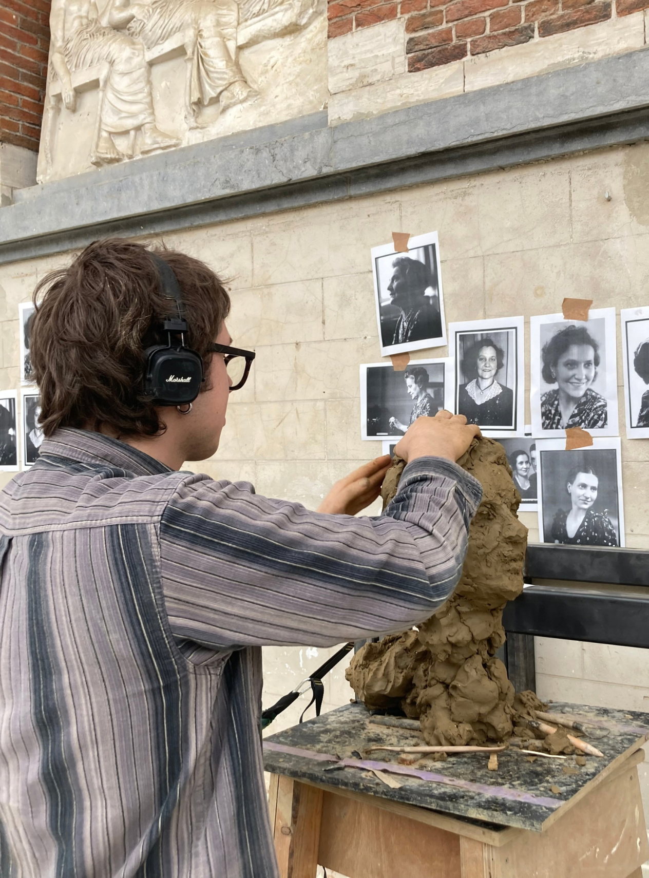 Foto van een jongen die geconcentreerd met koptelefoon op een beeld aan het sculpteren is. Op de muur voor hem hangen een aantal zwartwitfoto's van een vrouw.