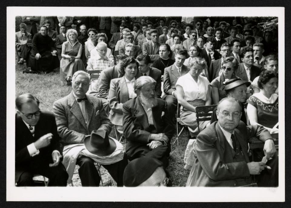Zwartwitfoto van een hele groep deftig geklede mensen op houten klapstoelen in een tuin
