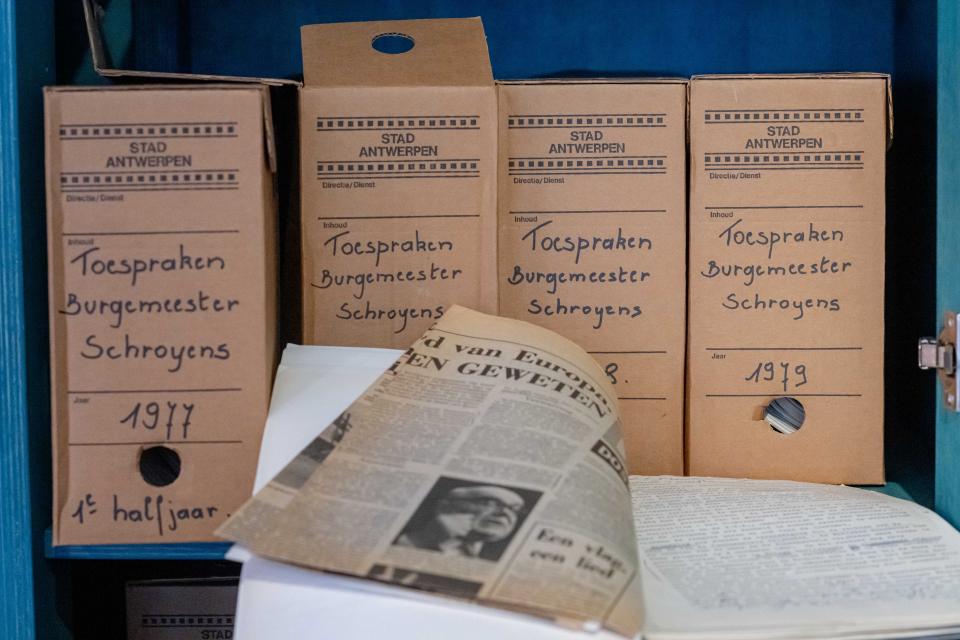 Vier bruine archiefdozen met het opschrift 'burgemeester Schroyens' staan in een kast geëtaleerd met daarvoor een openliggend schrift en een krantenknipsel.