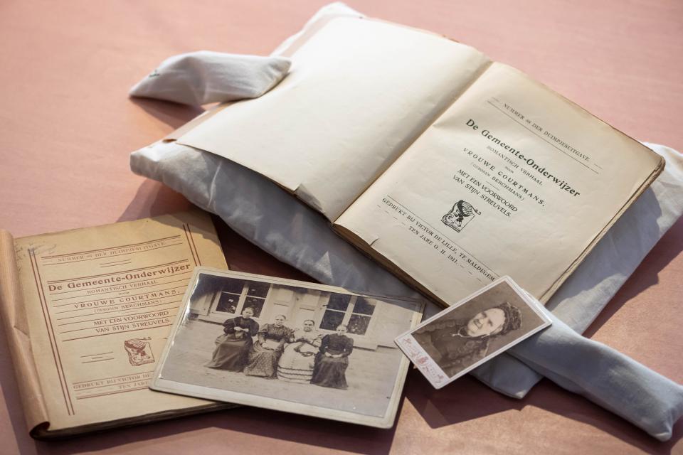 Archiefmateriaal van Johanna Courtmans-Berchmans op een tafel in het Letterenhuis, waaronder het opengeslagen boek 'De gemeente-onderwijzer' op de intropagina, een profielfoto in zwartwit van Courtmans-Berchmans, een foto waarop ze samen met drie andere vrouwen op een bankje zit.
