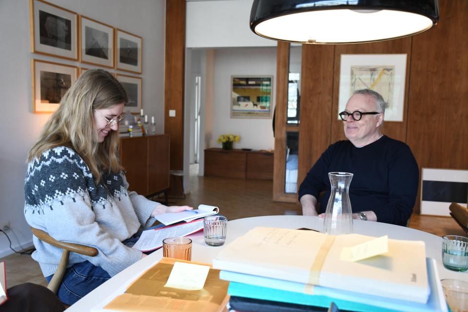 Een stagiaire van het Letterenhuis zit aan tafel met Willen van Zadelhoff in zijn woonkamer, terwijl ze notities maakt. Beiden lachen om iets wat net in het interview gezegd werd.