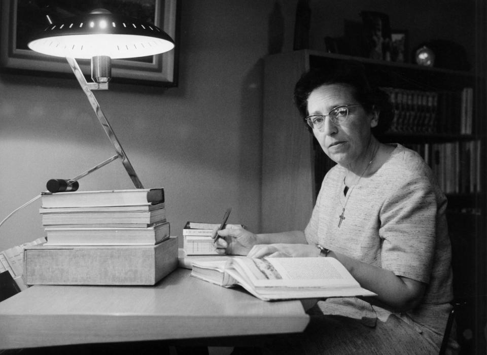 Zwartwit portretfoto van Maria Rosseels aan een bureau met een stapel dikke boeken voor zich en een brandende bureaulamp. Er ligt een boek open en ze neemt notities op een blocnote.