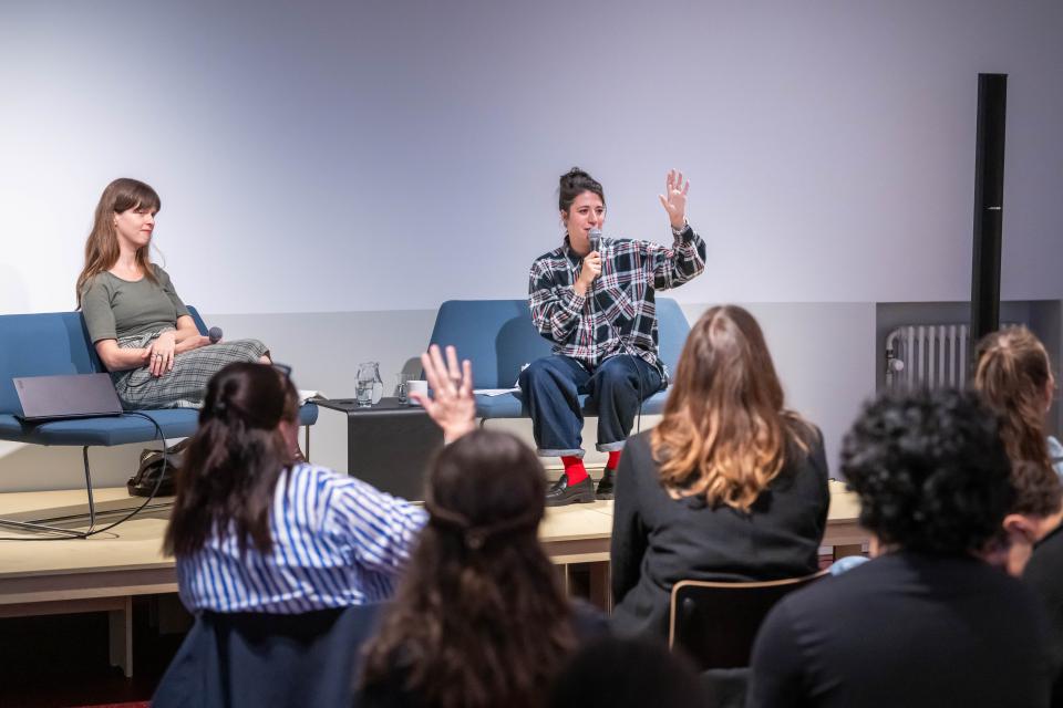 Dichter Sara Eelen en moderator Anke Verschueren zitten op podium in een salon. Voor het podium luisteren jongeren aandachtig naar het gesprek. Eén jongere steekt haar hand op.
