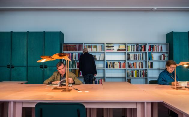 Een man en een vrouw zitten te lezen aan de tafels van de leeszaal terwijl een andere man de boeken in de bibliotheekkast bekijkt.