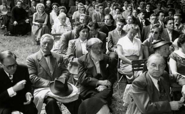 Zwartwitfoto van een hele groep deftig geklede mensen op houten klapstoelen in een tuin