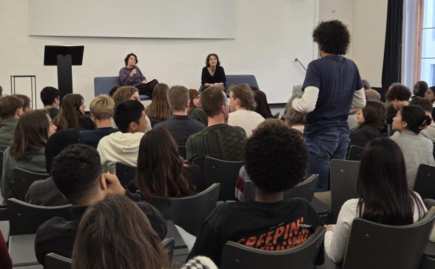 Een volle zaal scholieren luistert naar Hind Eljadid. Eén jongen staat recht om een vraag te stellen.