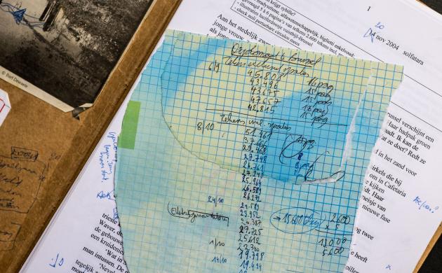 Erchiefstukken van Koen Peeters. Een bruine map ligt opengeslagen, op de linkerbinnenzijde kleeft een zwartwitfoto van een rivier met industriële gebouwen op de oever. Op de rechterzijde ligt een typoscript met correcties aangebracht in blauwe balpen en een geruit, afgescheurd blaadje met notatie van aantal tekens per hoofdstuk.