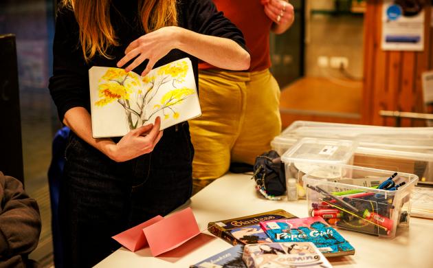 Iemand toont een zelfgemaakt zine. Op de tafel op de achtergrond liggen tijdschriften en knutselmateriaal.