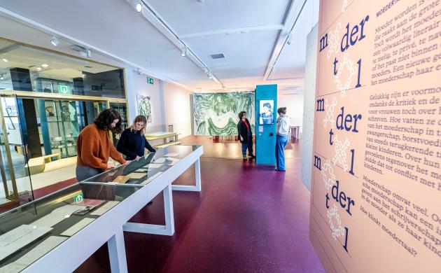In de exporuimte van het Letterenhuis staat de thema-expo Moeders opgesteld. 2 vrouwen bekijken de verhalen in een van de vitrinekasten, terwijl 2 andere vrouwen de video aan de zuil beluisteren.