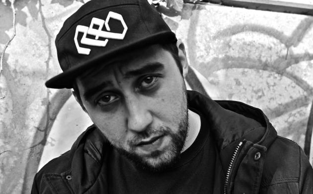 Portretfoto in zwartwit van Fatih De Vos voor een graffitimuur, met een zwarte sweater en zwarte pet. 