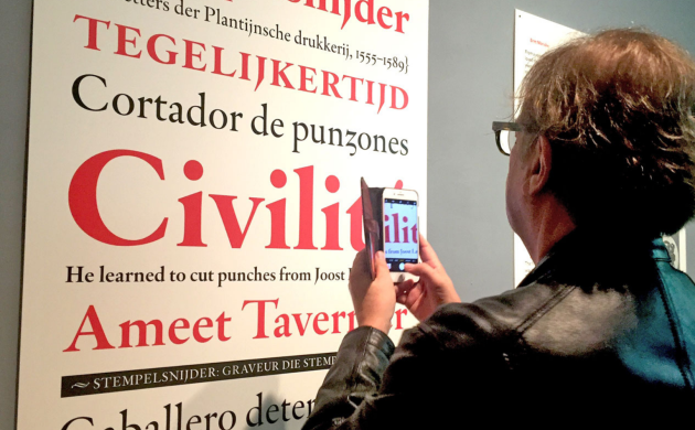 Aan een expomuur hangt een grote affiche met een lettertype in rood en zwart afgebeeld. Een man neemt er een foto van met zijn smartphone.