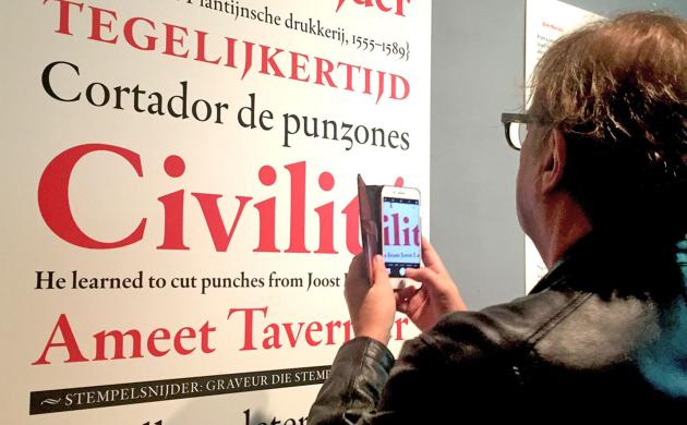 Een man met smartphone in de hand kijkt naar een grote print waarop in verschillende formaaten voorbeelden van een nieuw lettertype te zien zijn.