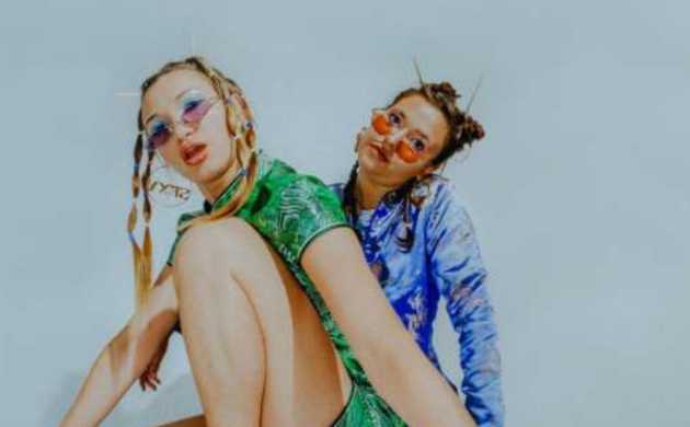 Mona Mina Leon en Zoë Heyninck gehurkt met zonnebrillen met gekleurde glazen en blinkende kleedjes aan.