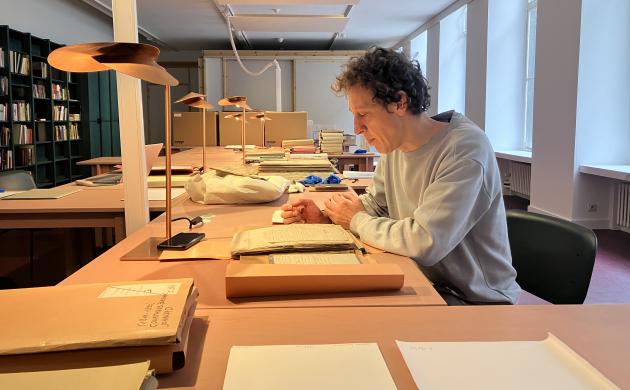 Tom Mariën zit aan een tafel in de leeszaal van het Letterenhuis, omringd door archiefmappen, en papieren.