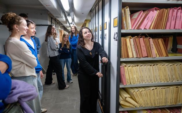 Een leerlinge draait aan het wiel om de archiefkasten in het depot te verplaatsen, terwijl de andere leerlingen van de klas lachend toekijken.