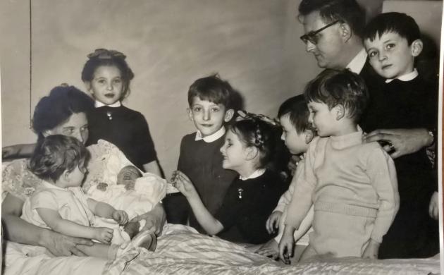 Een moeder houdt haar pasgeboren baby vast in een bed en errond staan de 7 andere kinderen en de vader