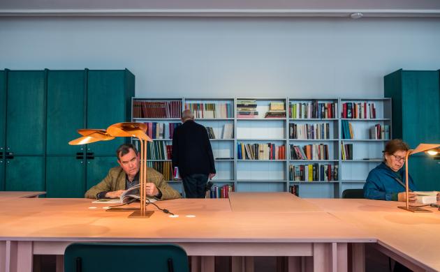 Een man en een vrouw zitten te lezen aan de tafels van de leeszaal terwijl een andere man de boeken in de bibliotheekkast bekijkt.