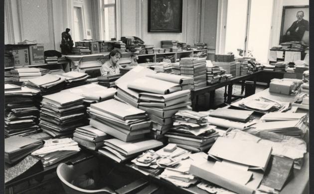 Alex Mallems in een bureauruimte omgeven door stapels documenten en mappen