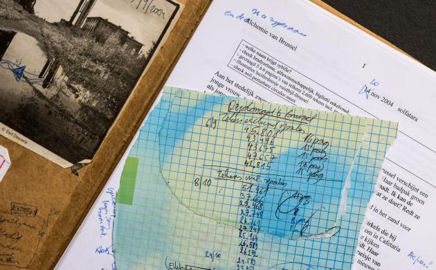 Erchiefstukken van Koen Peeters. Een bruine map ligt opengeslagen, op de linkerbinnenzijde kleeft een zwartwitfoto van een rivier met industriële gebouwen op de oever. Op de rechterzijde ligt een typoscript met correcties aangebracht in blauwe balpen en een geruit, afgescheurd blaadje met notatie van aantal tekens per hoofdstuk.