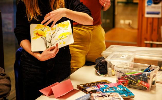 Iemand toont een zelfgemaakt zine. Op de tafel op de achtergrond liggen tijdschriften en knutselmateriaal.