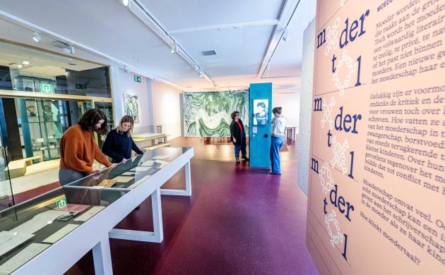 In de exporuimte van het Letterenhuis staat de thema-expo Moeders opgesteld. 2 vrouwen bekijken de verhalen in een van de vitrinekasten, terwijl 2 andere vrouwen de video aan de zuil beluisteren.