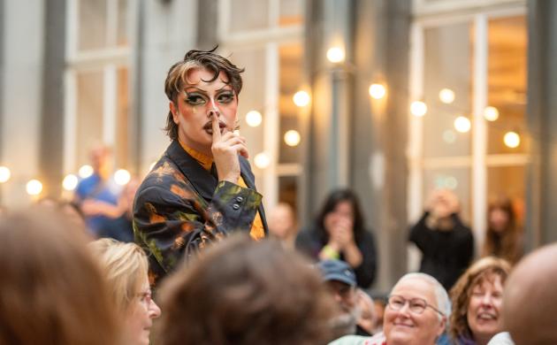 Een drag artiest staat op een podium en legt zijn vinger op zijn mond, als teken dat iedereen stil moet zijn. Blij gezichten kijken naar naar de show, op de achtergrond hangen brandende lichtjesslingers tegen de gevel van het Letterenhuis op de binnenkoer.
