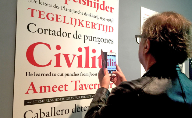 Aan een expomuur hangt een grote affiche met een lettertype in rood en zwart afgebeeld. Een man neemt er een foto van met zijn smartphone.