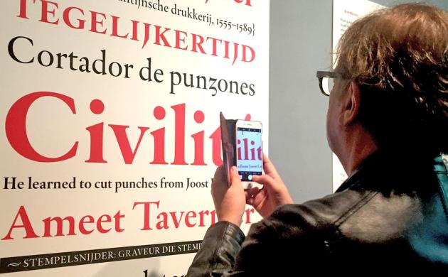 Een man met smartphone in de hand kijkt naar een grote print waarop in verschillende formaaten voorbeelden van een nieuw lettertype te zien zijn.