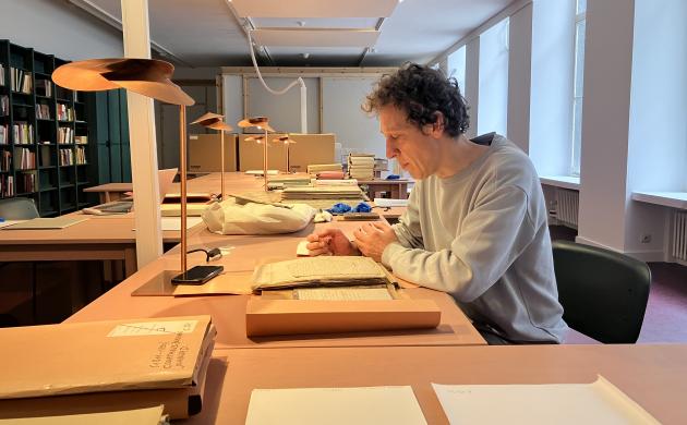 Tom Mariën zit aan een tafel in de leeszaal van het Letterenhuis, omringd door archiefmappen, en papieren.