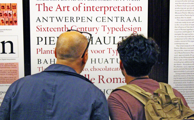 Twee mannen bekijken de expo Van letter tot boek. Ze kijken naar pancartes waarop lettertypes gedrukt staan in rood en zwart.
