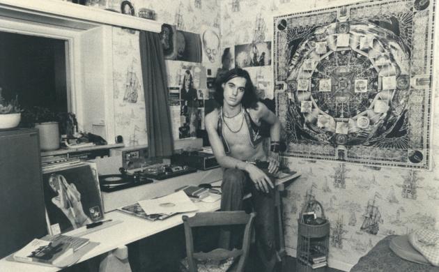 Jotie T'Hooft zit op een tafel in zijn werkkamer, waar LP's, een platenspeler, boeken op te zien zijn. Aan de muur hangt onder meer een mandala.