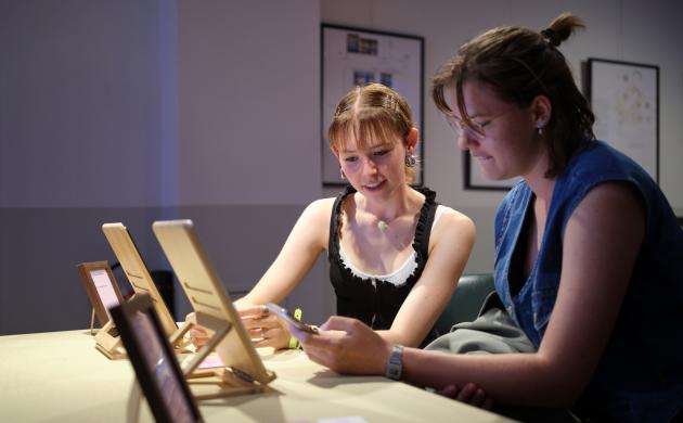 Twee jonge vrouwen, studentes, zitten aan een tafel in de studieruimte van het Letterenhuis en bekijken samen een archiefstuk. Op de tafel staan fotokaders en tablets.