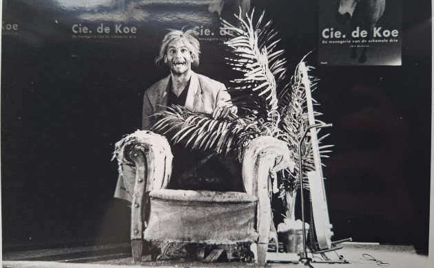 Zwartwitfoto van een man met een zeer brede lach en grote ogen die achter een versleten fauteuil staat. Op de achtergrond hangen affiches van De menagerie van de schamele drie.