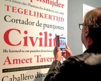 Een man met smartphone in de hand kijkt naar een grote print waarop in verschillende formaaten voorbeelden van een nieuw lettertype te zien zijn.